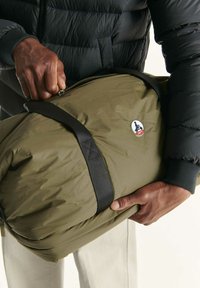 Una persona che indossa un giubbotto nero imbottito tiene una borsa da viaggio JOTT di colore verde oliva con manici neri e una cerniera.