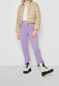 Beige uppblåst jacka med hög krage, lila sweatpants med vita sidremsor och krämfärgade klumpiga snörstövlar med tjock svart sula.