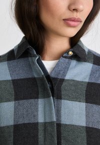 Camicia a quadri realizzata in tessuto morbido con un motivo nero, blu e verde, dotata di colletto con bottoni e un piccolo bottone di accentatura dorata.