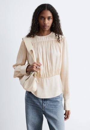 Młoda kobieta z kręconymi włosami nosi kremową plisowaną bluzkę, jasnoniebieskie dżinsy, kolczyki koła i trzyma beżową torbę na ramię.