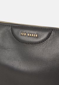 Zwarte leren tas met een zachte textuur, voorzien van een goudkleurig "TED BAKER" logo en een ritssluiting aan de bovenkant. Eenvoudig, strak ontwerp.