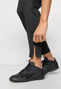 Μαύρα αθλητικά παπούτσια με ανάγλυφες επάνω επιφάνειες, χαμηλό προφίλ και λαστιχένιες σόλες με καρφιά, συνδυασμένα με μαύρο στενό παντελόνι που διαθέτει σχισμές στο τελείωμα.