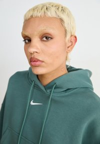 Πράσινο φούτερ από μαλακό ύφασμα με μπροστινή τσέπη, με λευκό λογότυπο Nike. Το μοντέλο έχει κοντά, ανοιχτόχρωμα μαλλιά και ένα μικρό τατουάζ με καρδιά.