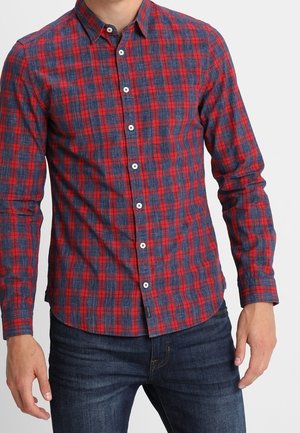 Mand iført en rød og mørkeblå ternet langærmet skjorte med knapper og mørkeblå jeans mod en ensfarvet baggrund.