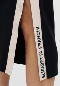 Μαύρο φόρεμα με πλαϊνή σχισμή και κρέμ λωρίδα. Η λωρίδα έχει κεντημένο το όνομα "ELISABETTA FRANCHI" με μαύρα γράμματα.