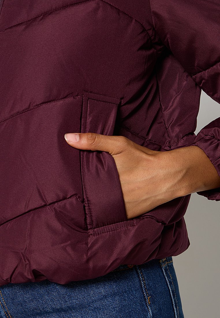 Burgunderfarbene Pufferjacke mit Seitentasche, gepolstertem Design und elastischen Bündchen. Das Material wirkt glatt und leicht glänzend.