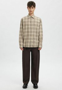 Beež ruuduline nööbitav särk pikkade varrukatega, lõdva istuvusega ja klassikalise kraega, kombineeritud tumedate sirge lõikega pükste ja mustade kingadega.