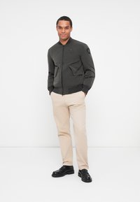Giubbotto bomber verde scuro con polsini a costine, due tasche frontali con patta e una toppa circolare sulla spalla. Indossato con pantaloni beige chiaro e scarpe nere.