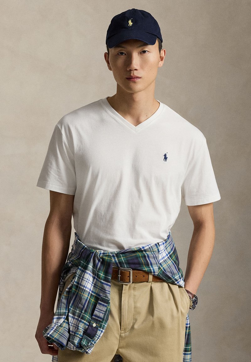 White cotton v-neck t-shirt featuring a small blue logo. Beige khaki pants with a brown belt. Navy cap with yellow logo, plaid shirt tied at the waist.
