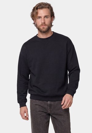 Mand med lysbrunt hår og skæg iført en simpel sort sweatshirt og mørkegrå jeans, stående foran en ensfarvet hvid baggrund.