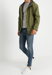 Vattenavvisande jacka i olivgrönt med justerbar huva, kombinerad med en ljusgrå skjorta och smala blå jeans, accessoriserad med vita sneakers.