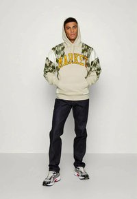 Smėlinis džemperis su "MARKET" geltonomis raidėmis, kamufliažo rašto rankovės, derinamas su tamsiomis džinsomis ir daugiaspalviais sportbačiais.