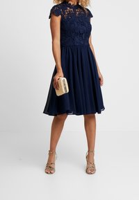 Robe bleu marine avec un corps en dentelle, des manches courtes et une taille ajustée. Jupe en chiffon évasée. Associée à une petite pochette tissée et des sandales métalliques à lanières.