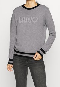 Nainen, jolla on päällään harmaa neulepaita mustilla yksityiskohdilla ja "LIU JO" -logo, yhdistettynä tummanharmaisiin farkkuihin, poseeraa yksi käsi vyötäröllä.