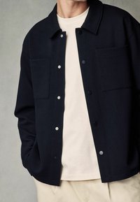 Tmavě modrá texturovaná bunda se dvěma náprsními kapsami, zapínáním na knoflíky a rozšířeným límcem, nošená přes světle béžovou košili.