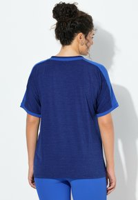 Kobieta z kręconymi włosami związanymi w kok, ubrana w niebieską koszulkę z krótkim rękawem i dopasowane niebieskie spodnie, zwrócona tyłem na białym tle.