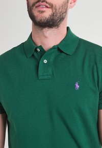 Grön polo t-shirt i texturerad tyg med klassisk krage. Har en tvåknappslå och en liten lila logotyp på bröstet.