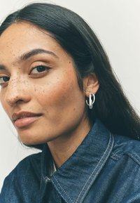Orecchino a cerchio in argento con un design attorcigliato, indossato da una persona con capelli scuri. La giacca di jeans ha un colletto strutturato e cuciture visibili.
