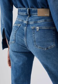 Inimese lähivõte, kes kannab siniseid teksapükse ja sobivat teksasärki, näidates tagataskuid ja pruunist nahast kaubamärgiga silti vööjoonel.