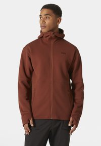 Chaqueta marrón con cremallera fabricada en un tejido texturizado, con un cuello alto, bolsillos laterales y orificios para los pulgares. Presenta un logotipo en el pecho.