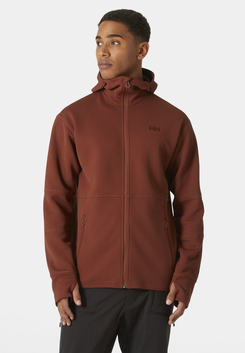 Chaqueta marrón con cremallera fabricada en un tejido texturizado, con un cuello alto, bolsillos laterales y orificios para los pulgares. Presenta un logotipo en el pecho.