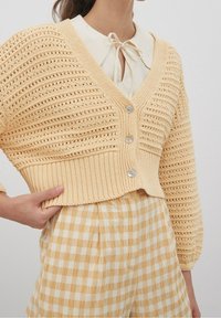 Frau trägt eine beige gestrickte, verkürzte Strickjacke mit silbernen Knöpfen über einer cremefarbenen Bluse und einem gelben karierten Rock.