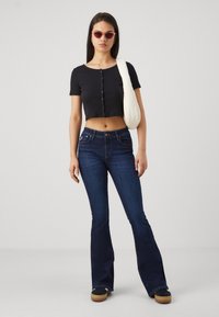 Svart kort knappad topp, mörkblå utsvängda jeans och svarta platåskor; modellen bär en krämfärgad axelväska och rosa solglasögon.