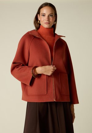 Woman wearing rust-colored zip-up jacket over matching turtleneck with dark skirt, posing against plain beige background.