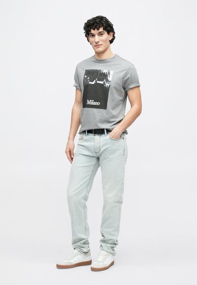 Γκρι t-shirt με μαύρο γραφικό σχέδιο με την επιγραφή «Milano», συνδυασμένο με ανοιχτό μπλε τζιν και λευκά αθλητικά παπούτσια με καφέ σόλες.