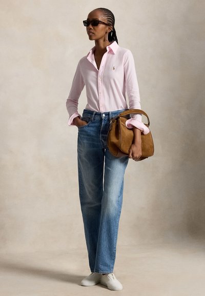 Donna con capelli intrecciati e occhiali da sole, indossa una camicia rosa con bottoni, jeans blu, sneakers bianche, tiene una borsa di pelle marrone.