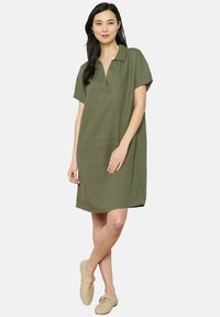 Vestido de lino verde oliva con cuello, mangas cortas y aberturas en los laterales. La modelo lleva calzado perforado beige y está de pie sobre un fondo plano.