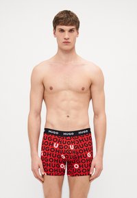 Boxer-briefs en coton rouge avec une ceinture noire ornée de texte "HUGO" répété en différentes tailles. Design ajusté.