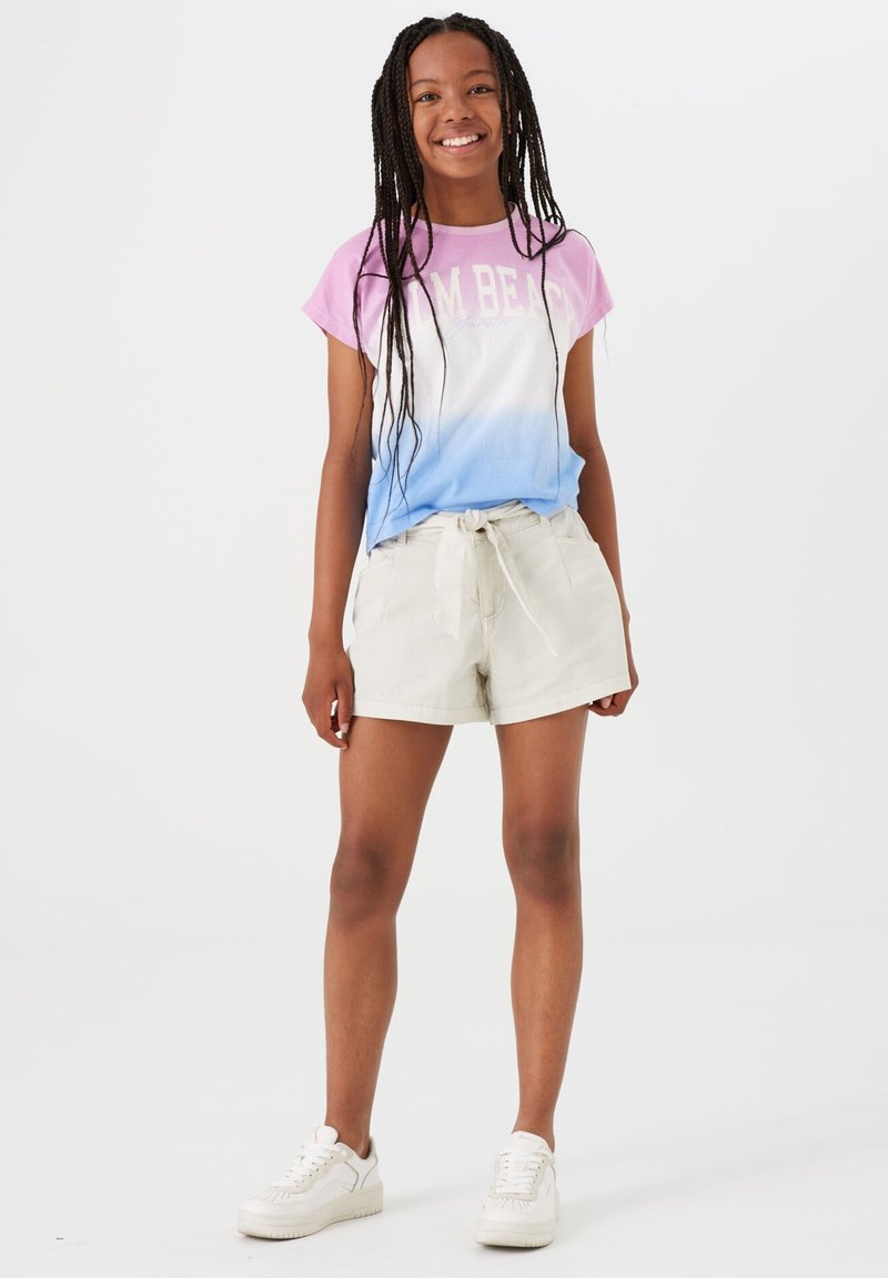 Het kortste mouw shirt met een gradient heeft een roze naar blauwe ombre, met de tekst "Palm Beach". Gecombineerd met lichtbeige shorts en witte sneakers.