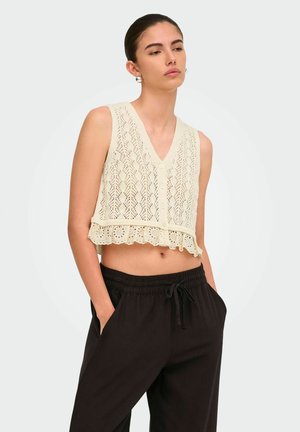 Tânără purtând un top scurt croșetat, de culoare crem, fără mâneci, cu nasturi, asortat cu pantaloni largi negri cu șnur, stând cu mâinile în buzunare.