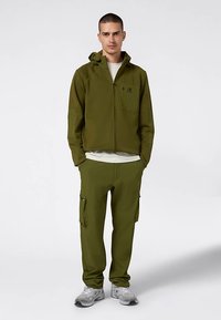 Olijfgroene jas met rits en capuchon, voorzak; bijpassende cargobroek met zijzakken; lichtgewicht stof; gedragen met een wit T-shirt en sneakers.