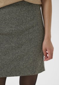 Jupe en mélange de laine de couleur vert foncé avec un motif crème moucheté, à coupe droite et fente latérale, associée à un haut beige et des collants noirs.