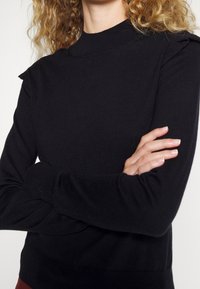 Osoba o kręconych blond włosach nosi czarny sweter z długim rękawem i golfem, z założonymi na piersi złożonymi ramionami.