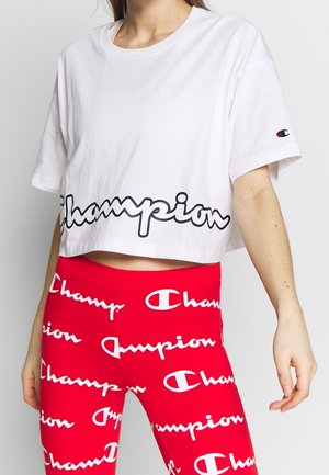 Osoba nosząca biały krótki t-shirt Champion oraz czerwone legginsy z powtarzającymi się białymi logo i napisem Champion.