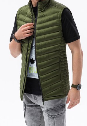 Homme portant un gilet matelassé vert olive ouvert sur un t-shirt graphique noir et un pantalon cargo gris clair.