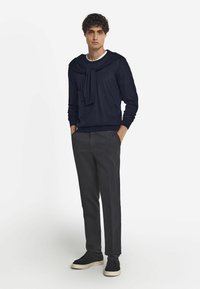 Marinblå stickad tröja med draperad frontdetalj, kombinerad med grå byxor och svarta sneakers med vita sulor, placerad mot en enfärgad bakgrund.