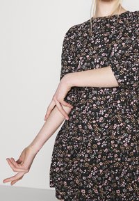 Vrouw past de mouw van een zwarte jurk met een klein roze en oranje bloemenpatroon aan, tegen een effen lichte achtergrond.