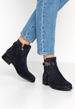 Bottes bottines en daim bleu marine avec fermeture éclair latérale et sangle noire ornée d'une boucle dorée "TH", portées avec un jean bleu retroussé.