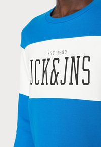 Modrý a bílý svetr s kulatým výstřihem, na přední straně má výrazný černý text "JCK&JNS" a "EST. 1990". Vyrobeno z měkké bavlny.