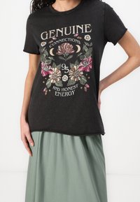 Vrouw draagt een zwart T-shirt met bloemenontwerp en de tekst "Echte Verbindingen 95 en Eerlijke Energie" in combinatie met een lichtgroene rok.