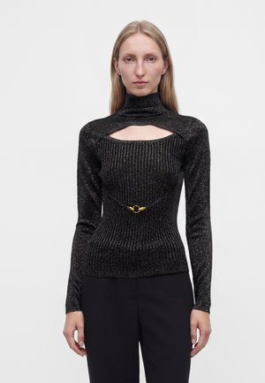 Femme aux longs cheveux blonds porte un top côtelé noir pailleté avec une découpe haute au col et un détail boucle dorée, associé à un pantalon noir.