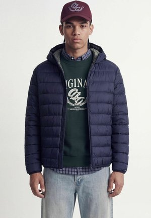 Homme portant une casquette bordeaux, une doudoune bleu marine, un sweat-shirt vert foncé avec un texte, une chemise à carreaux en dessous et un jean bleu clair.