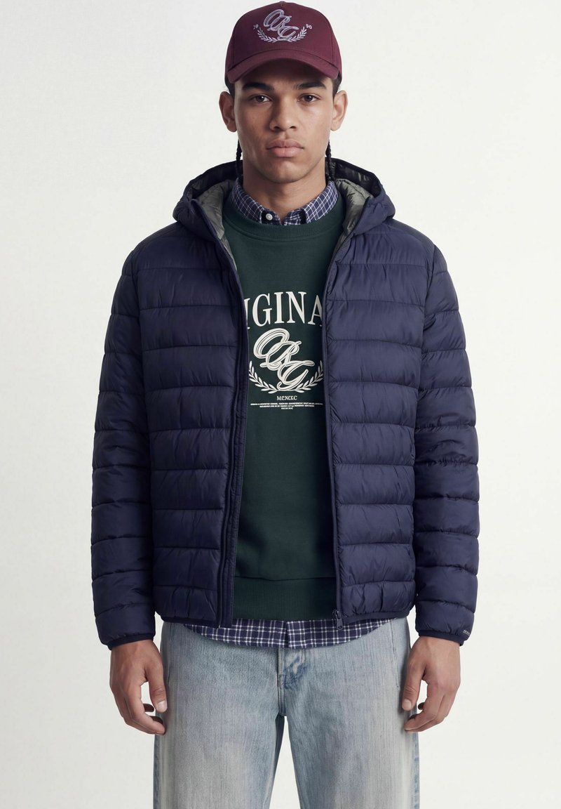 Homme portant une casquette bordeaux, une doudoune bleu marine, un sweat-shirt vert foncé avec un texte, une chemise à carreaux en dessous et un jean bleu clair.