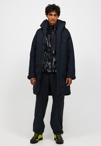 Svart pufferjacka med pälsfodrad huva över en mönstrad svart och grå zip-up tröja, tillsammans med mörka byxor och kängor med gröna detaljer.