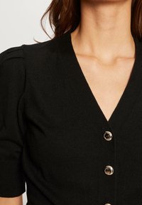 Schwarze gestrickte Strickjacke mit V-Ausschnitt, kurzen Puffärmeln und drei runden goldfarbenen Knöpfen an der Vorderseite. Glatte Textur, figurschnitt.