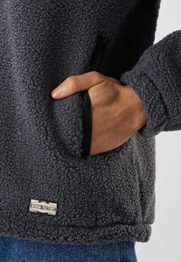 Main dans la poche d’une veste polaire texturée gris foncé avec une étiquette Denim Factory, associée à un jean bleu.